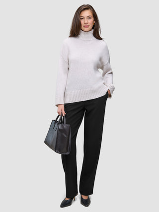 En kvinna klädd i en krämfärgad tröja med hög hals och svarta Women GO2 Pants Straight, som håller en svart handväska.
