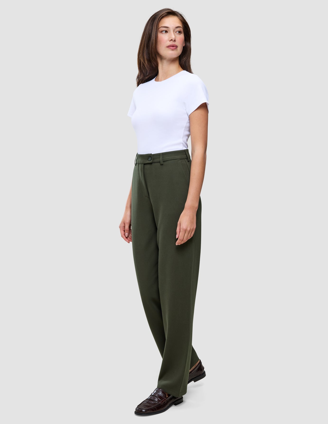 En kvinna bär en vit t-shirt instoppad i ett par högmidjade Women GO2 Pants Straight i färgen Dusty Olive samt mörka loafers.