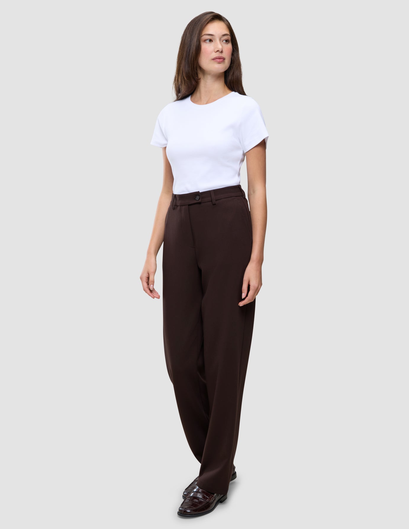 En kvinna bär en vit t-shirt och byxor av modellen Women GO2 Pants Straight Mocha i en rak, brun modell, tillsammans med mörka loafers.