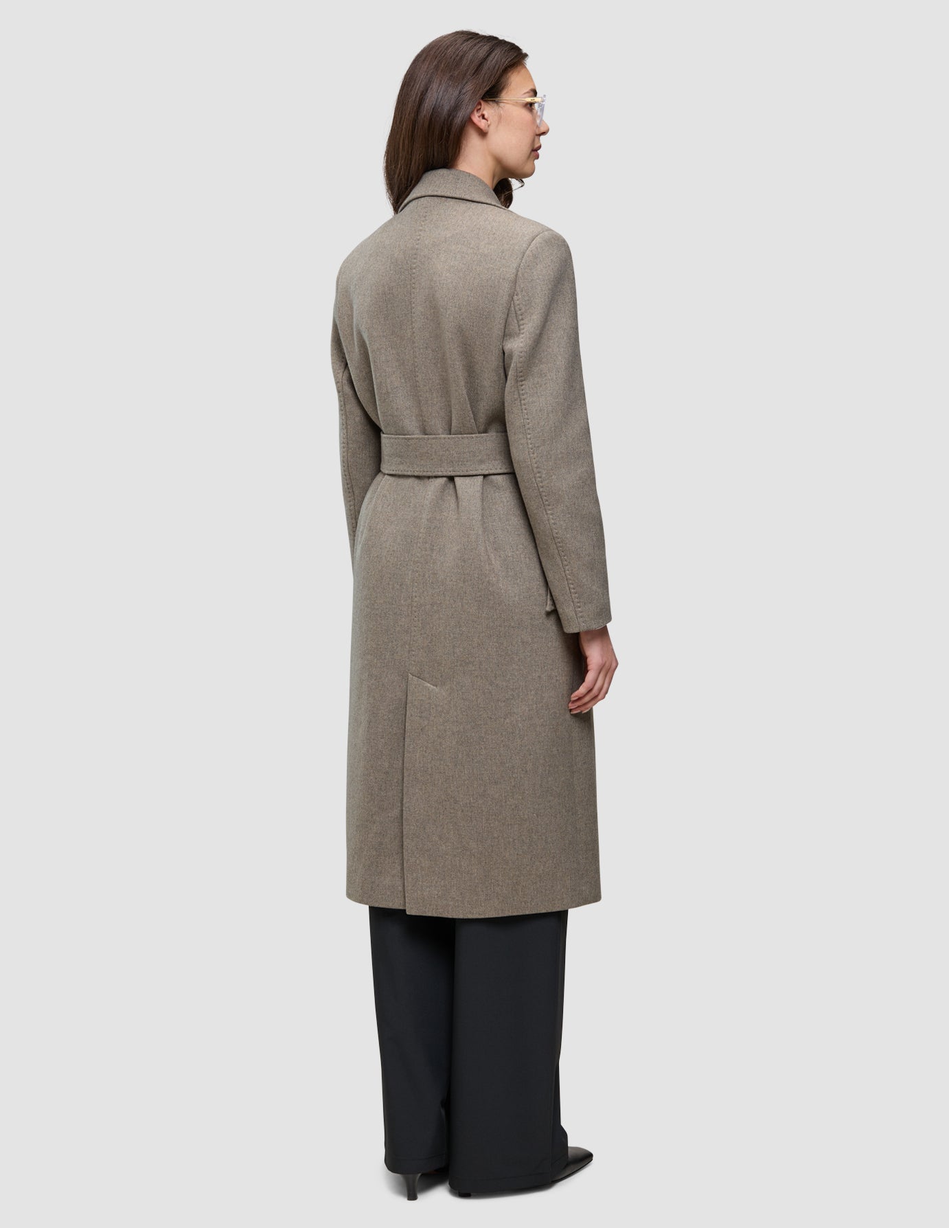En kvinna sedd bakifrån, klädd i en gråbeige damrock i ull, i kombination med svarta byxor i minimalistisk stil.