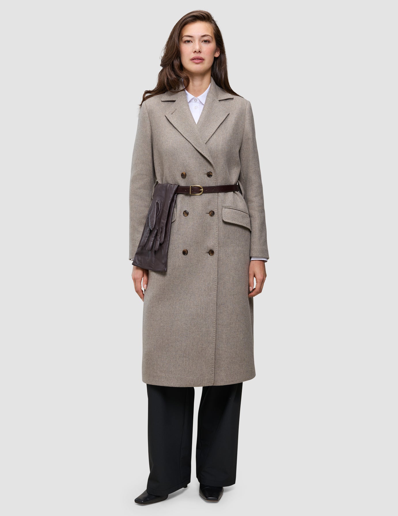 En kvinna bär en vinterklädsel bestående av en elegant beige ullkappa med ett brunt bälte och läderhandskar.
