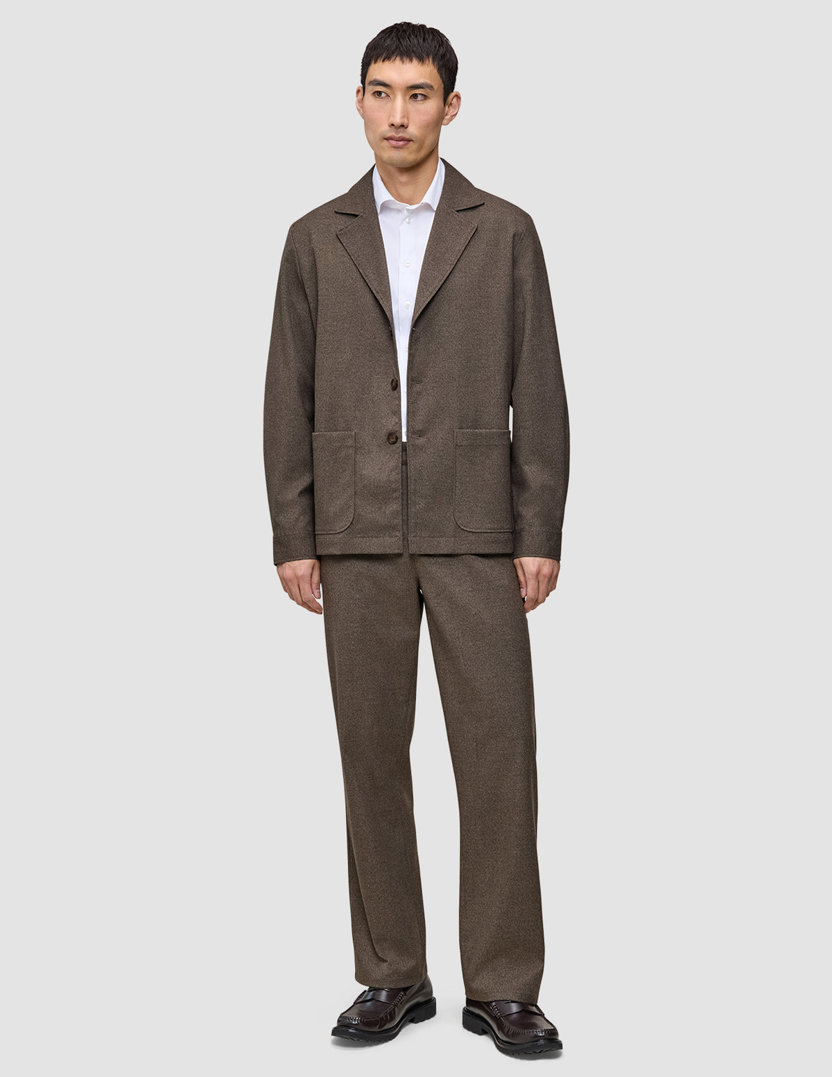 En man klädd i en mörkbeige overshirt från Atelier, en vit skjorta med krage, matchande byxor och mörkbruna loafers.