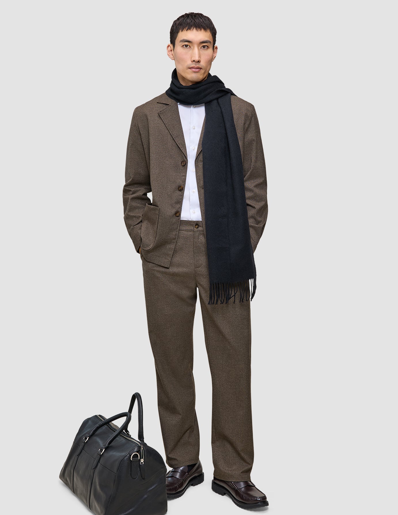 En man bär en brun, rutig Atelier Overshirt Dark Beige i twill, en vit skjorta och en svart halsduk bredvid en svart läderväska.