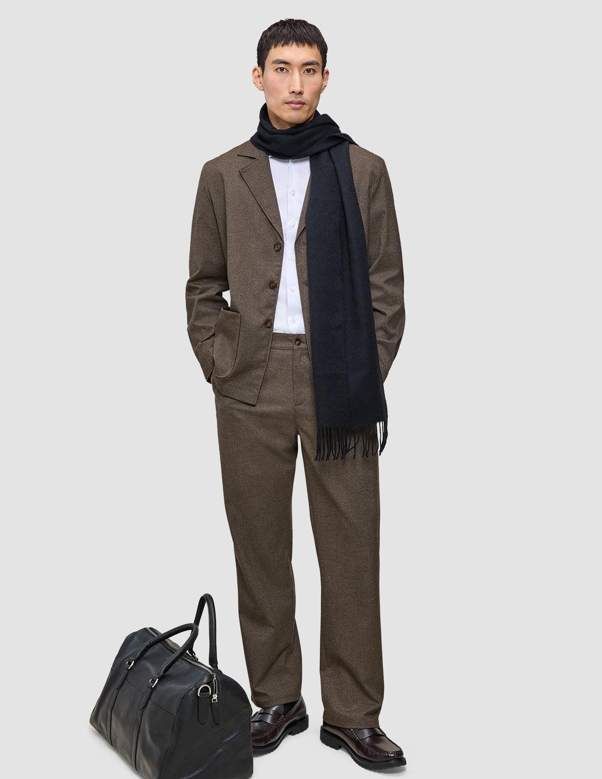 En man bär en brun, rutig Atelier Overshirt Dark Beige i twill, en vit skjorta och en svart halsduk bredvid en svart läderväska.