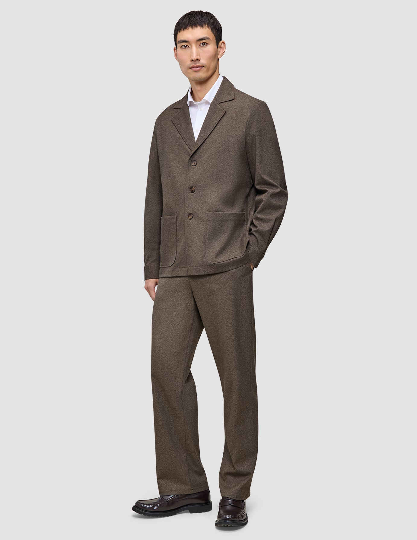 En man klädd i en mörkbeige overshirt från Atelier, vit skjorta och matchande byxor samt mörka loafers.