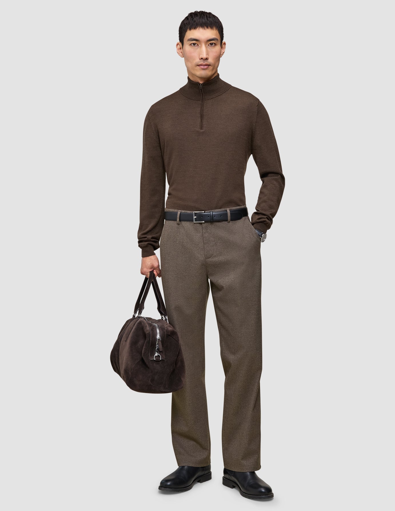 En man klädd i en brun tröja med kvartslång dragkedja och byxor av modellen Atelier Pants Relaxed Dark Beige, som håller i en mörk väska.