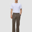 En man i en vit t-shirt, byxor av modellen Atelier Pants Relaxed i mörkbeige och svarta läderskor.