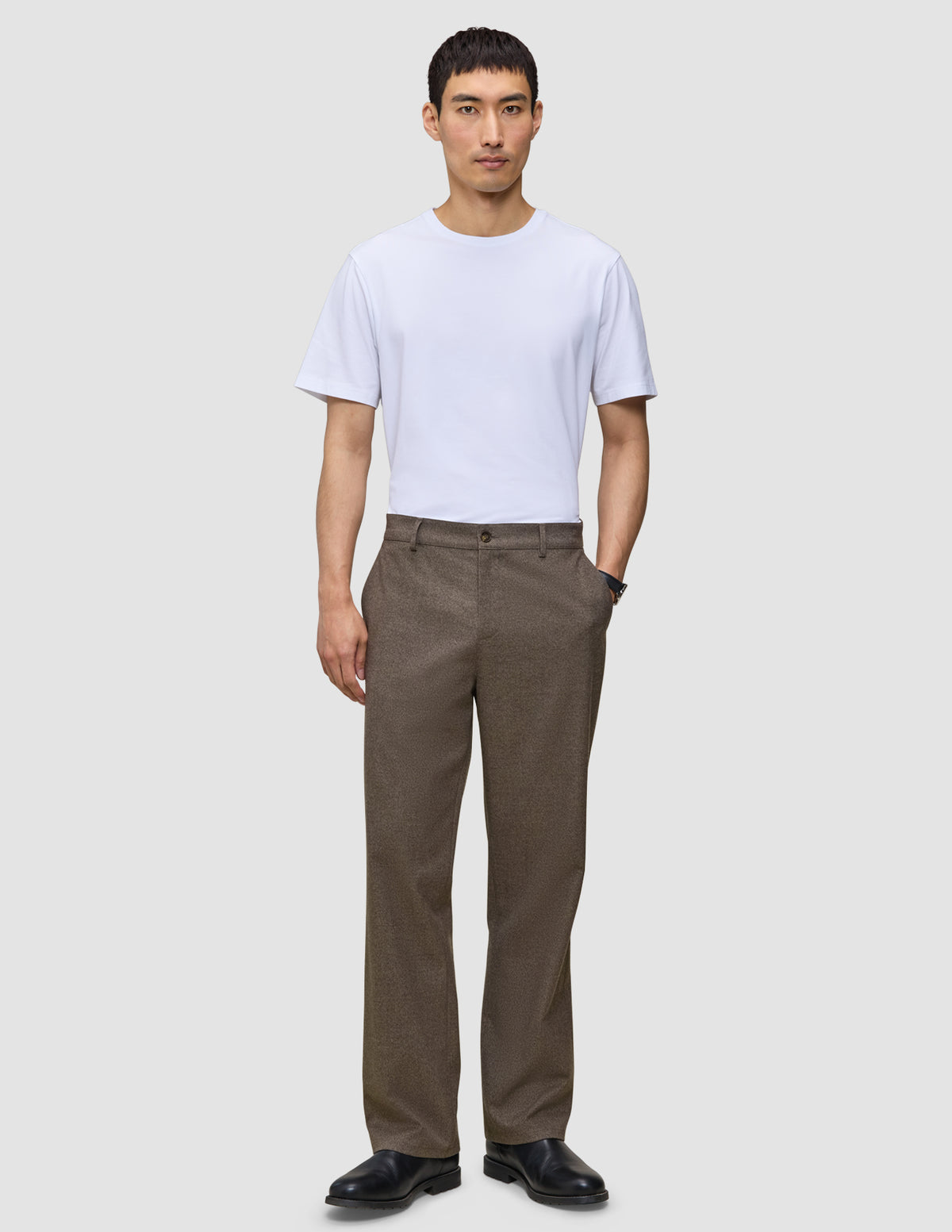 En man i en vit t-shirt, byxor av modellen Atelier Pants Relaxed i mörkbeige och svarta läderskor.