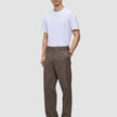 En man i en vit t-shirt, byxor av modellen Atelier Pants Relaxed i mörkbeige och svarta kostymskor.