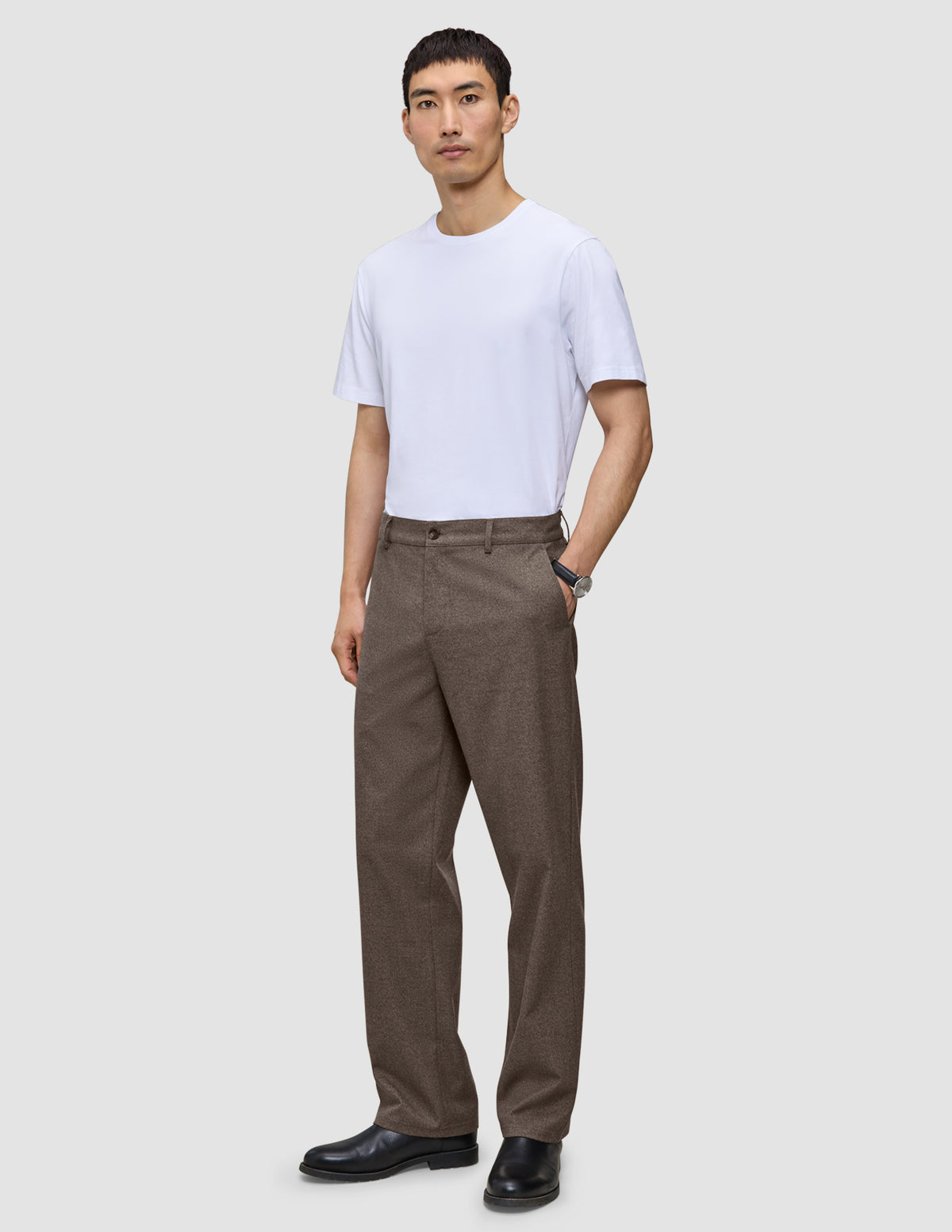 En man i en vit t-shirt, byxor av modellen Atelier Pants Relaxed i mörkbeige och svarta kostymskor.