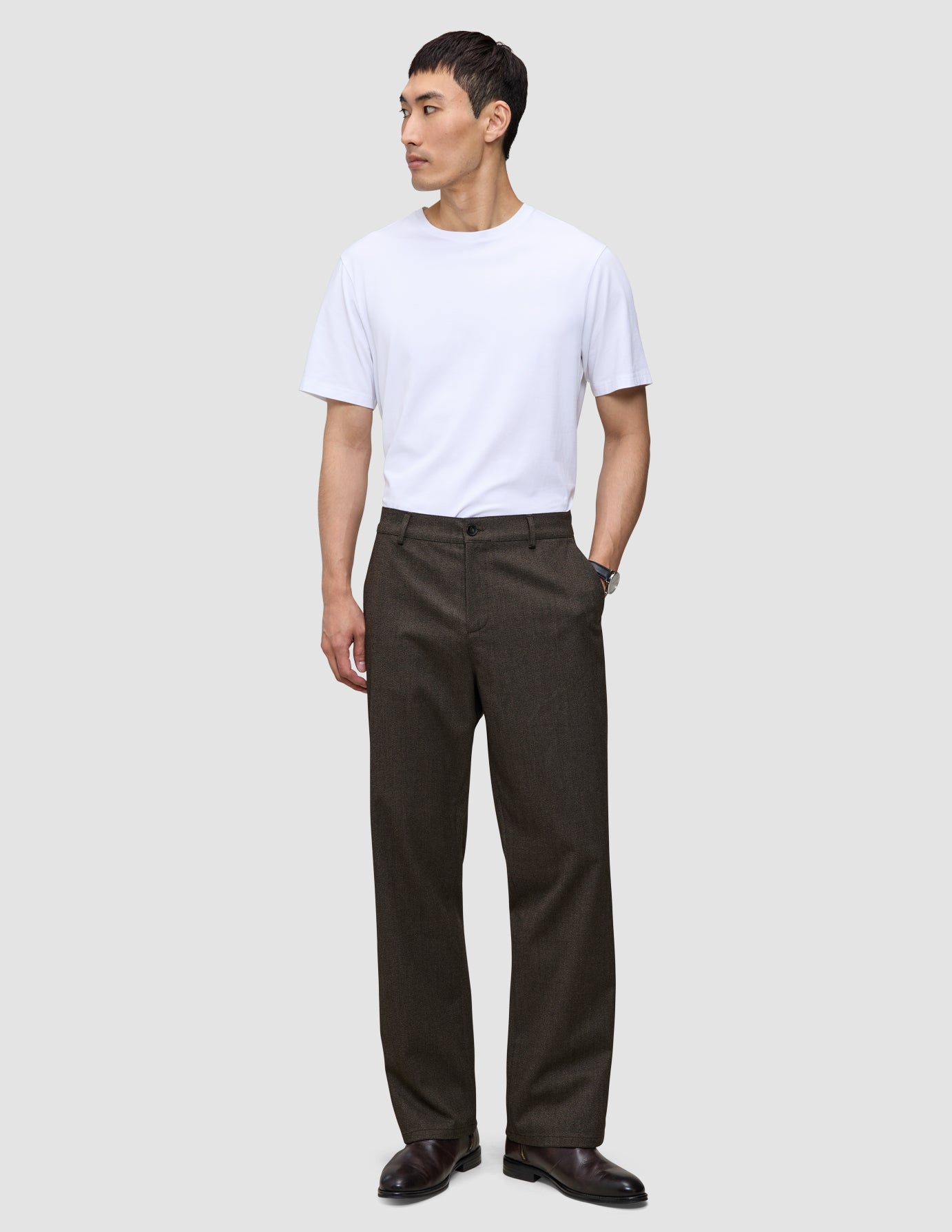 En person i en vit t-shirt, byxor av modellen Atelier Pants Relaxed i mörkbrunt och mörka läderskor.