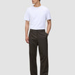 En person i en vit t-shirt, byxor av modellen Atelier Pants Relaxed i mörkbrunt och mörka läderskor.