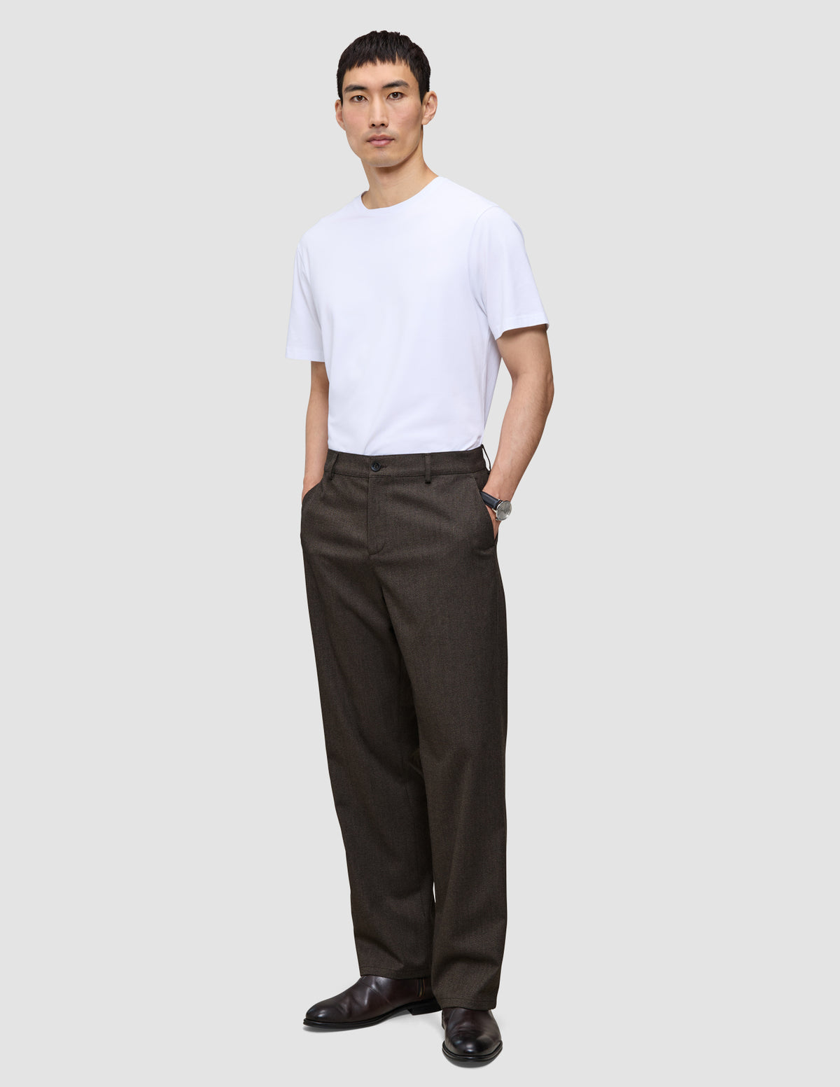 En person i en vit t-shirt och mörkbruna Atelier Pants Relaxed med avslappnad passform, med händerna i fickorna.