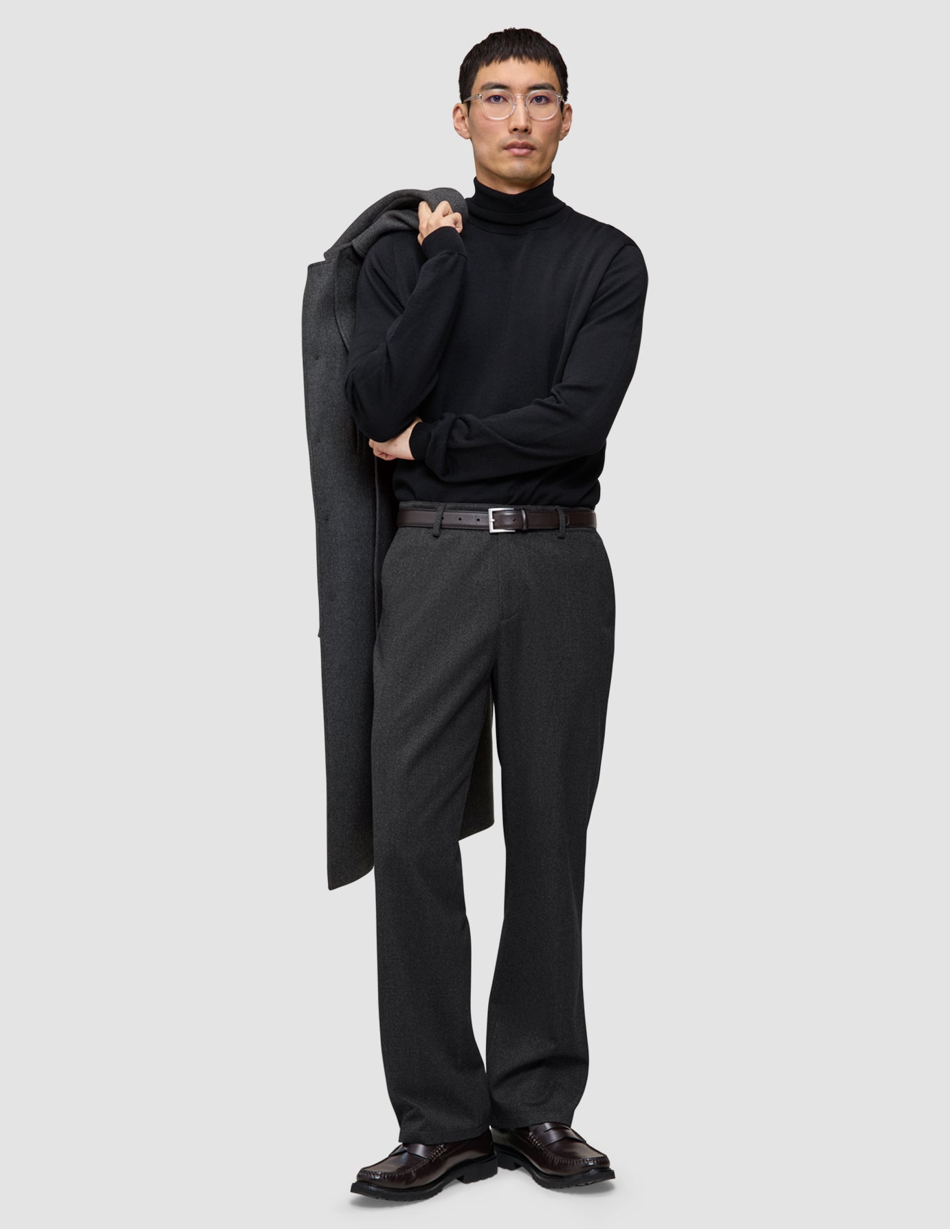 En man i svart polotröja och mörkgråa Atelier Pants Relaxed, med en kappa slängd över axeln i skandinavisk stil.