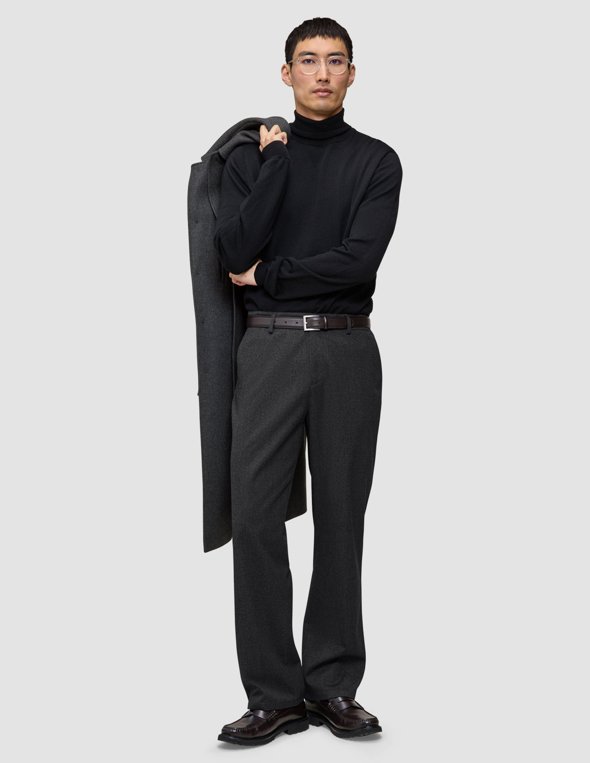 En man i svart polotröja och mörkgråa Atelier Pants Relaxed, med en kappa slängd över axeln i skandinavisk stil.