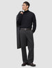 En man i svart polotröja och ett par Atelier Pants Relaxed i mörkgrått med avslappnad passform, med en kappa över axeln.