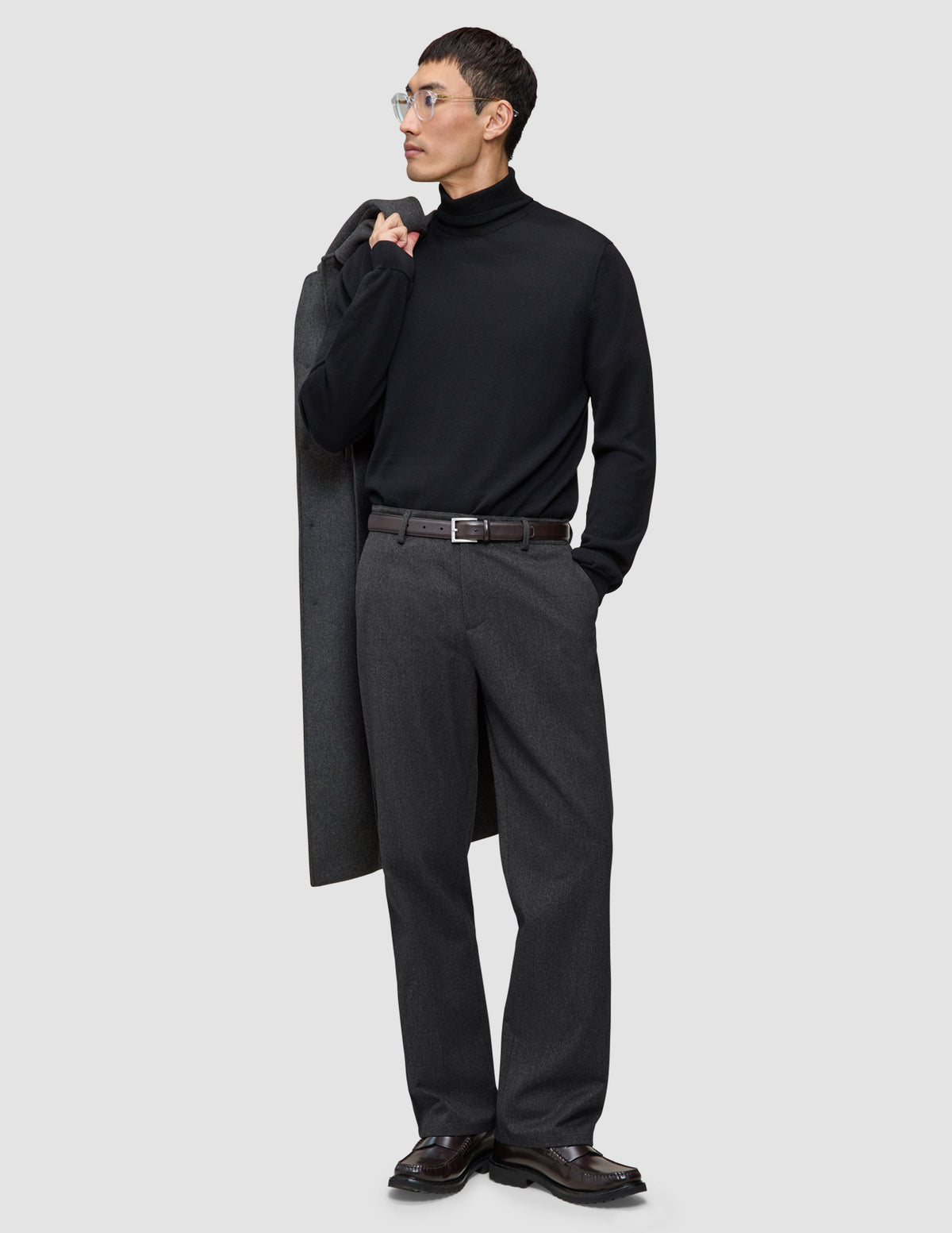 En man i svart polotröja och ett par Atelier Pants Relaxed i mörkgrått med avslappnad passform, med en kappa över axeln.