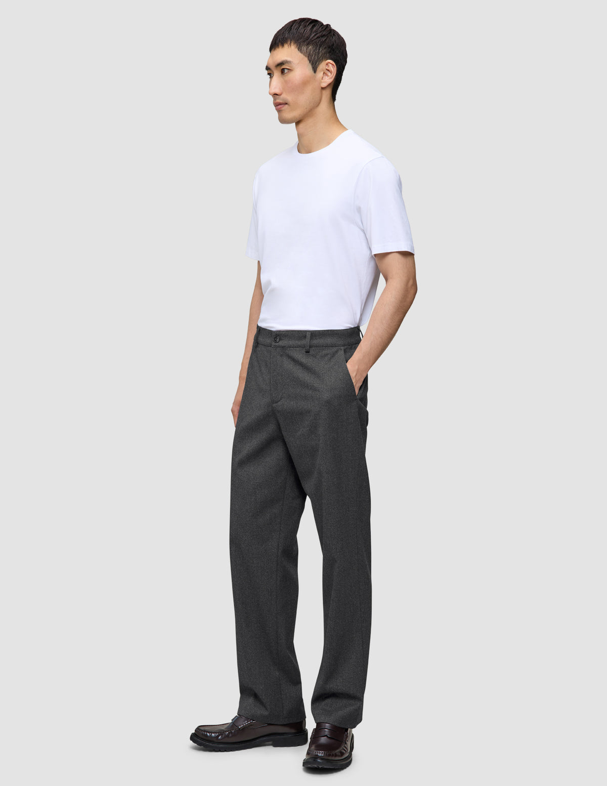 En person bär en vit t-shirt, byxor av modellen Atelier Pants Relaxed i mörkgrått och mörka loafers.