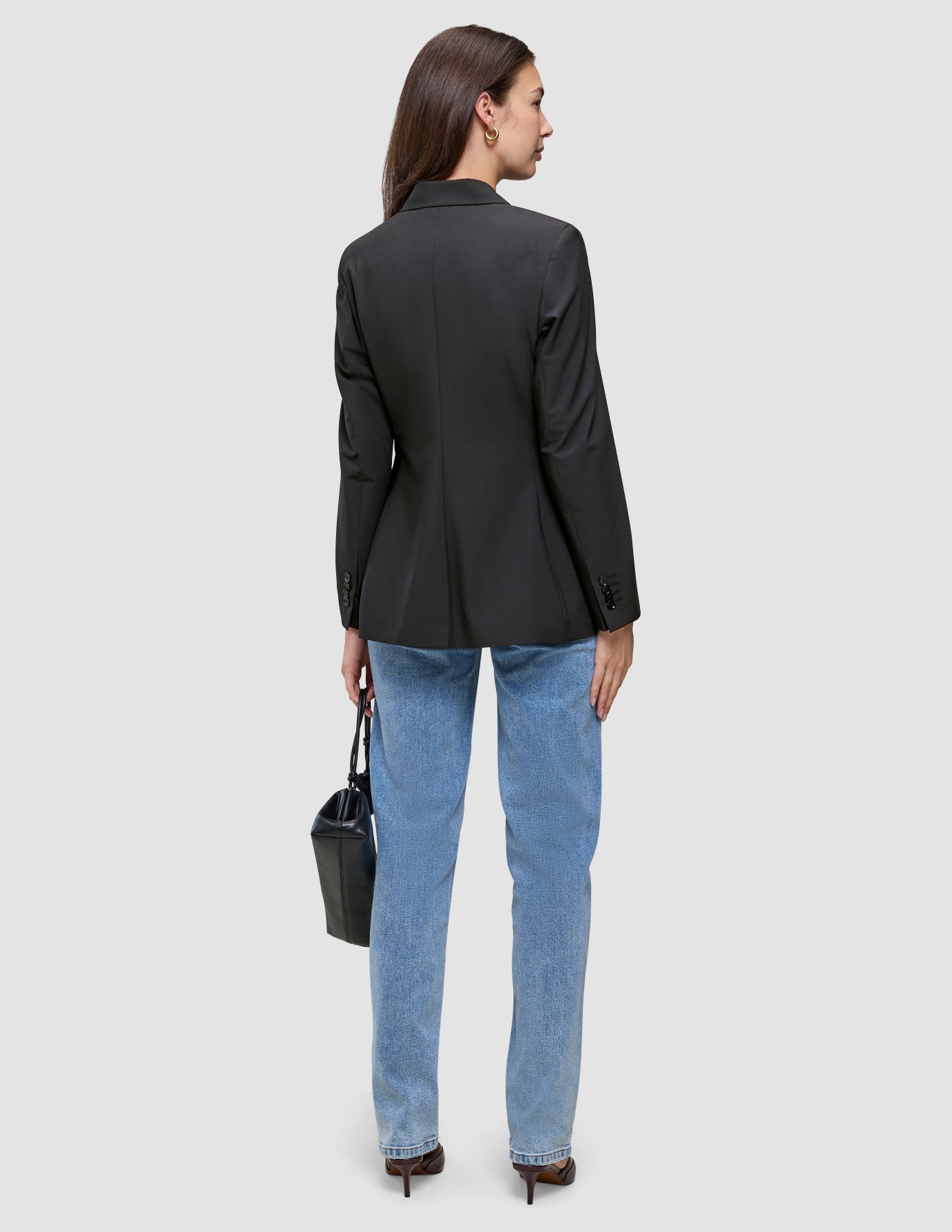 En person sedd bakifrån, klädd i en svart Avira-kavaj för damer, ljusblå jeans och högklackade skor, som håller i en svart handväska.