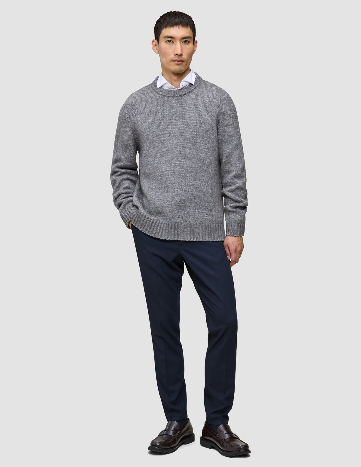 En man bär en tjock, mjuk ulltröja med rund hals i färgen Glacier Grey över en vit skjorta, marinblå byxor och mörka loafers.