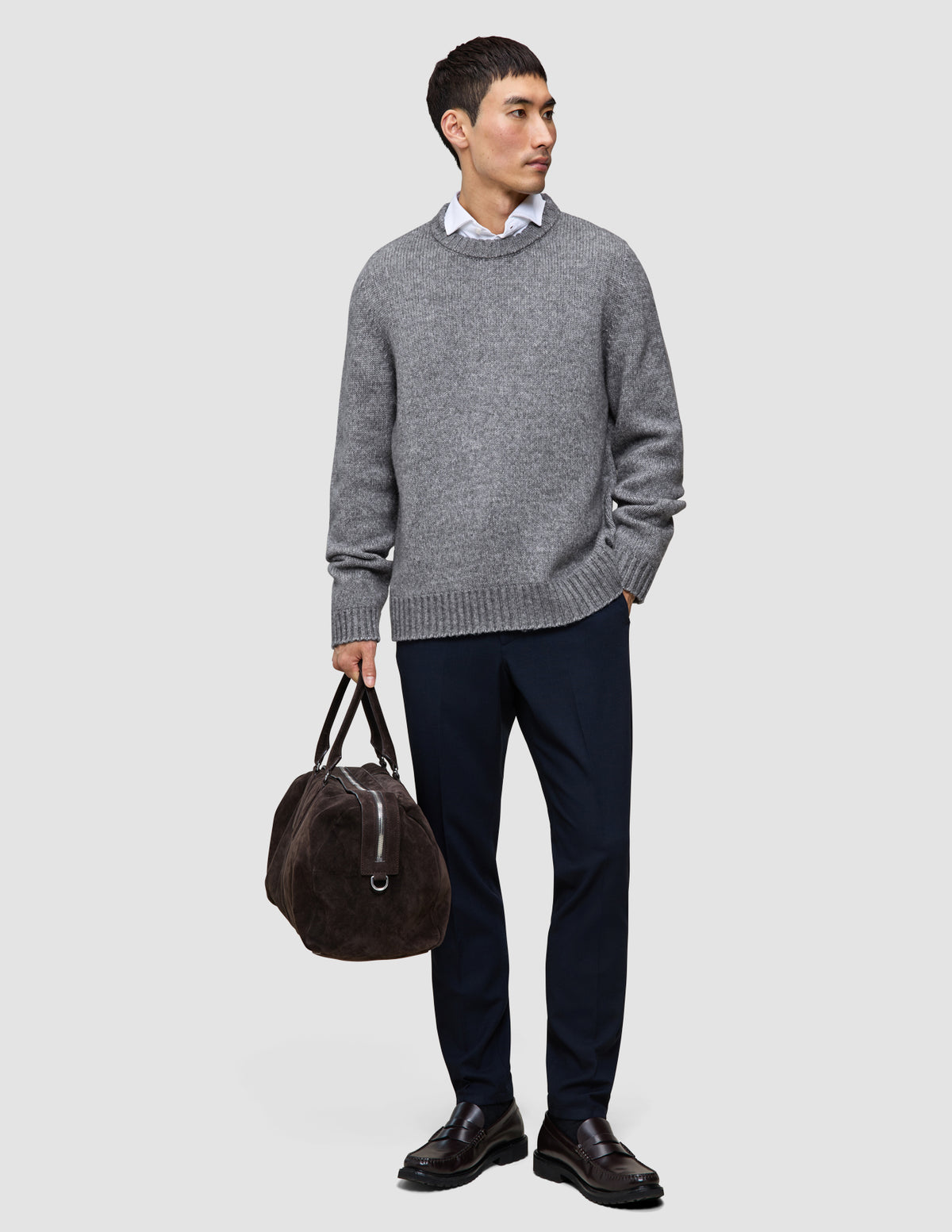 En man bär en grå, tjock och mjuk ulltröja med rund hals i färgen Glacier Grey över en skjorta med krage, mörka byxor och loafers.