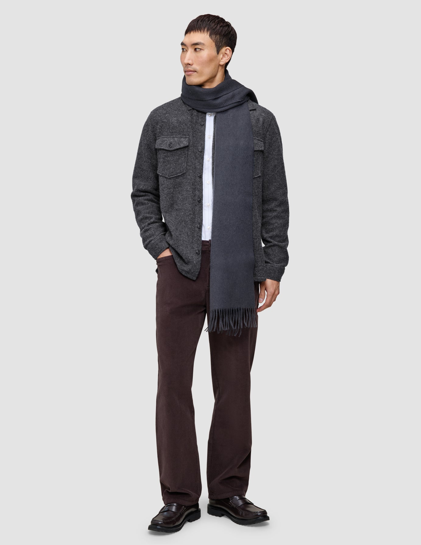 En man bär en dubbelskiktad yttertröja i ull, modell Regular, mörkgrå, över en vit T-shirt, tillsammans med en grå halsduk, bruna byxor och loafers.