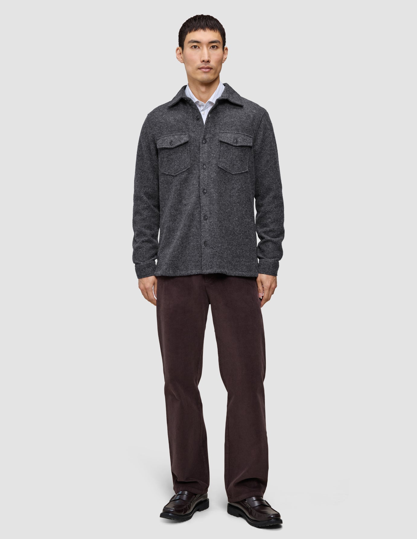 En man bär en dubbellagad yttertröja i ull, modell Regular, mörkgrå, över en vit skjorta, tillsammans med mörka byxor och loafers.
