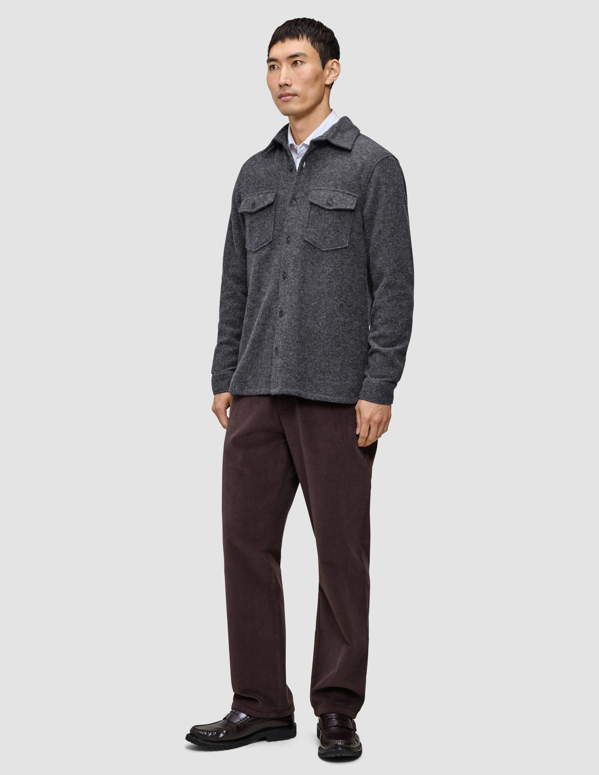 En man bär en dubbelskiktad yttertröja i ull, modell Regular, mörkgrå, över en vit skjorta, mörka byxor och loafers.