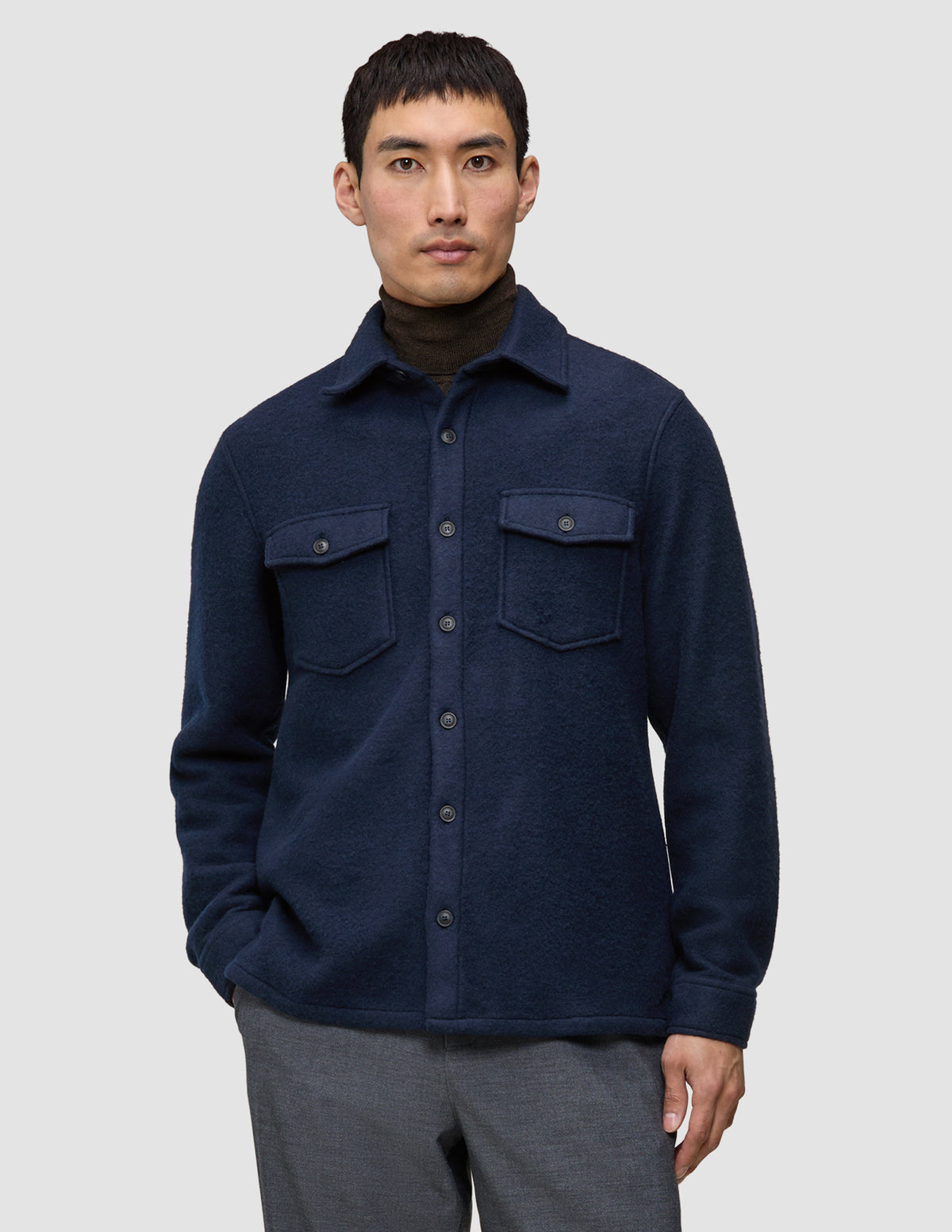 En man i en dubbelskiktad yttertröja i ull, modell Regular, mörk marinblå, över en brun polotröja, vilket återspeglar en minimalistisk design.