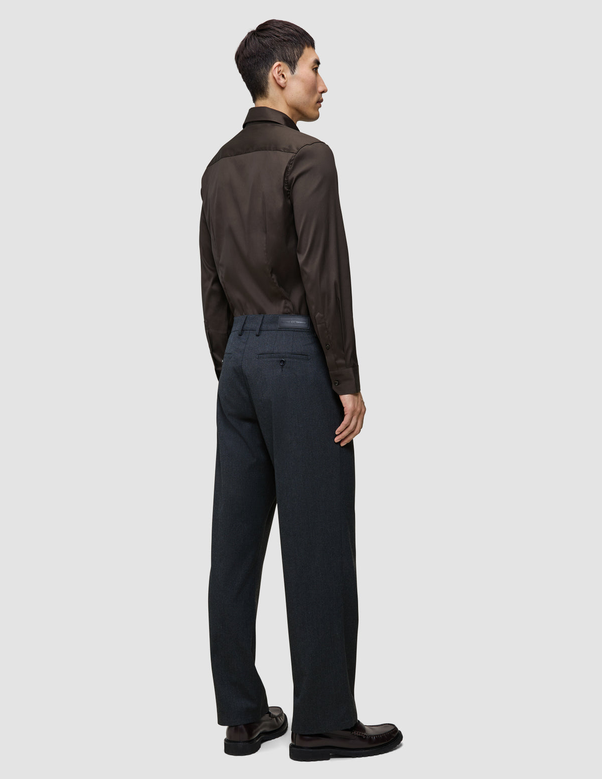 En man sedd bakifrån, klädd i en mörkbrun, figursydd skjorta med hög krage, kolgrå byxor och mörka loafers.
