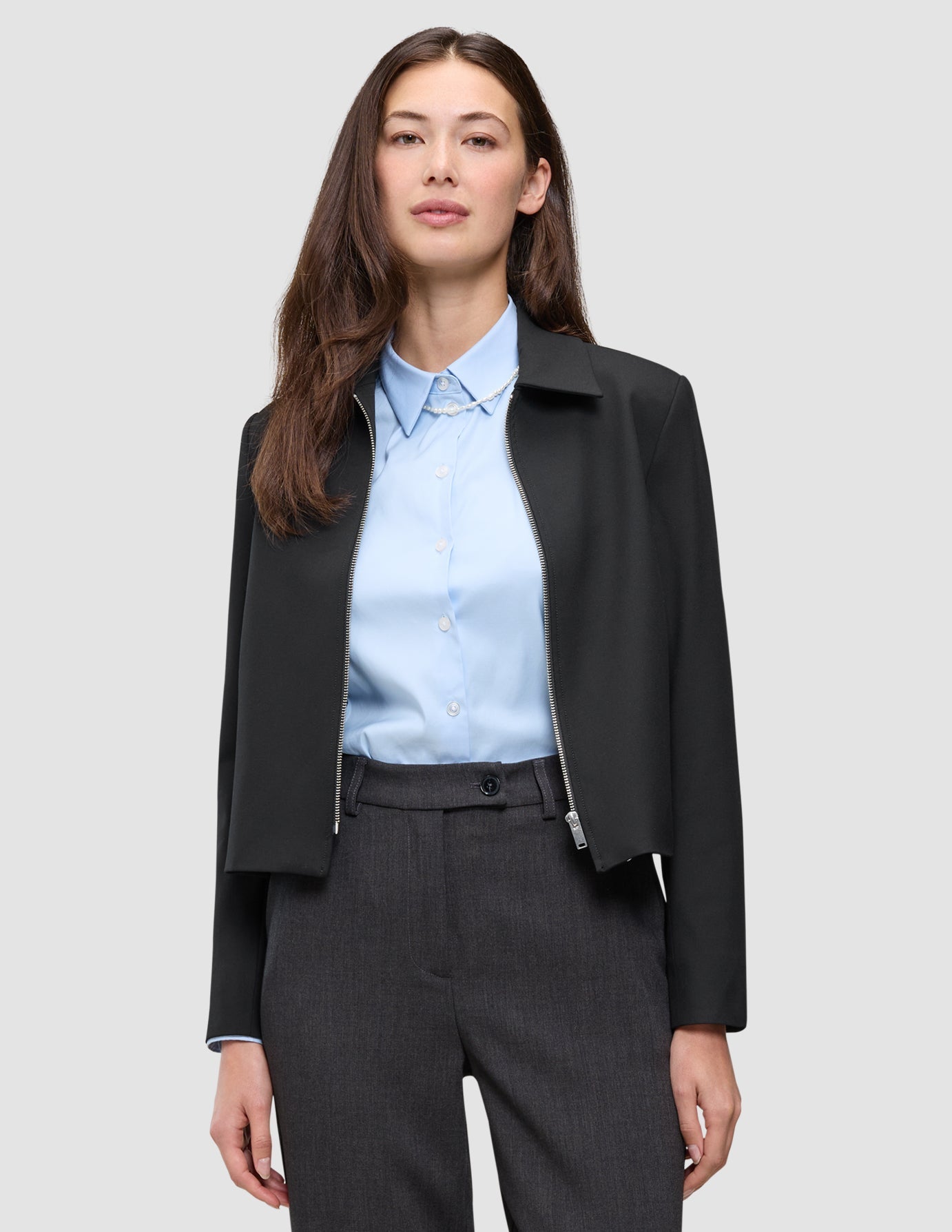 En kvinna bär en svart Essential Short Zip Jacket över en ljusblå skjorta med knappar och grå byxor.