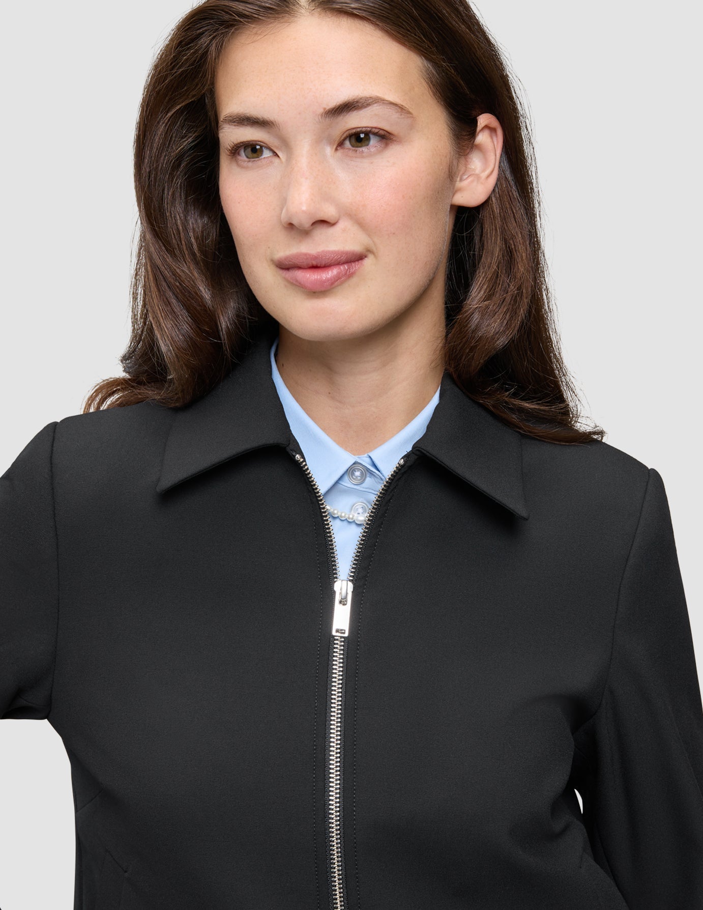 En person som bär en minimalistisk Essential Short Zip Jacket Black över en ljusblå skjorta med knappar.