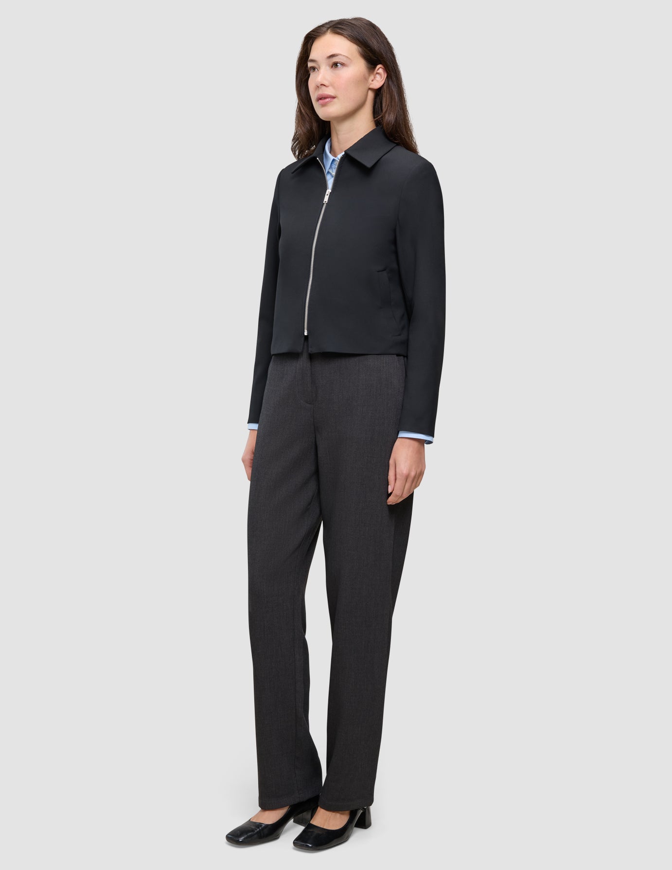 En kvinna bär en svart Essential Short Zip Jacket över en ljusblå skjorta med krage och mörkgrå byxor.