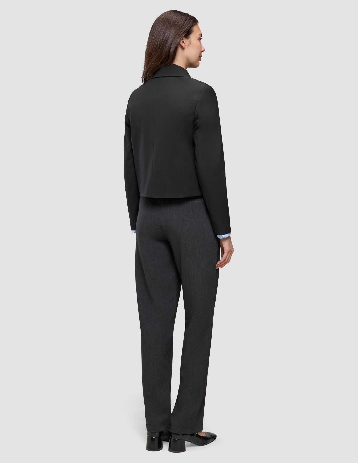 En kvinna sedd bakifrån, klädd i en svart Essential Short Zip Jacket över en ljusblå skjorta med krage och mörkgrå byxor.
