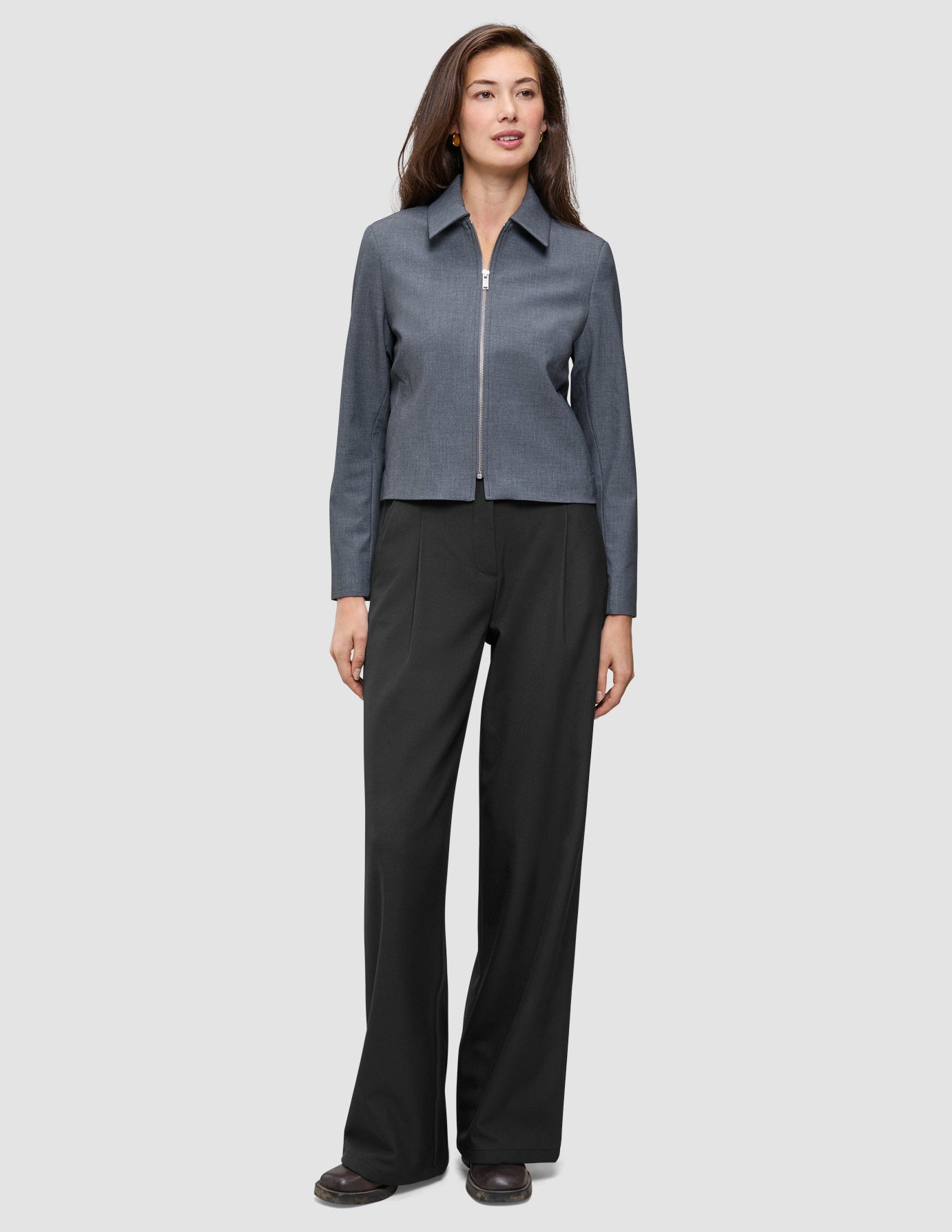 En kvinna klädd i en minimalistisk Essential Short Zip Jacket i mörkgrå melange och svarta byxor.