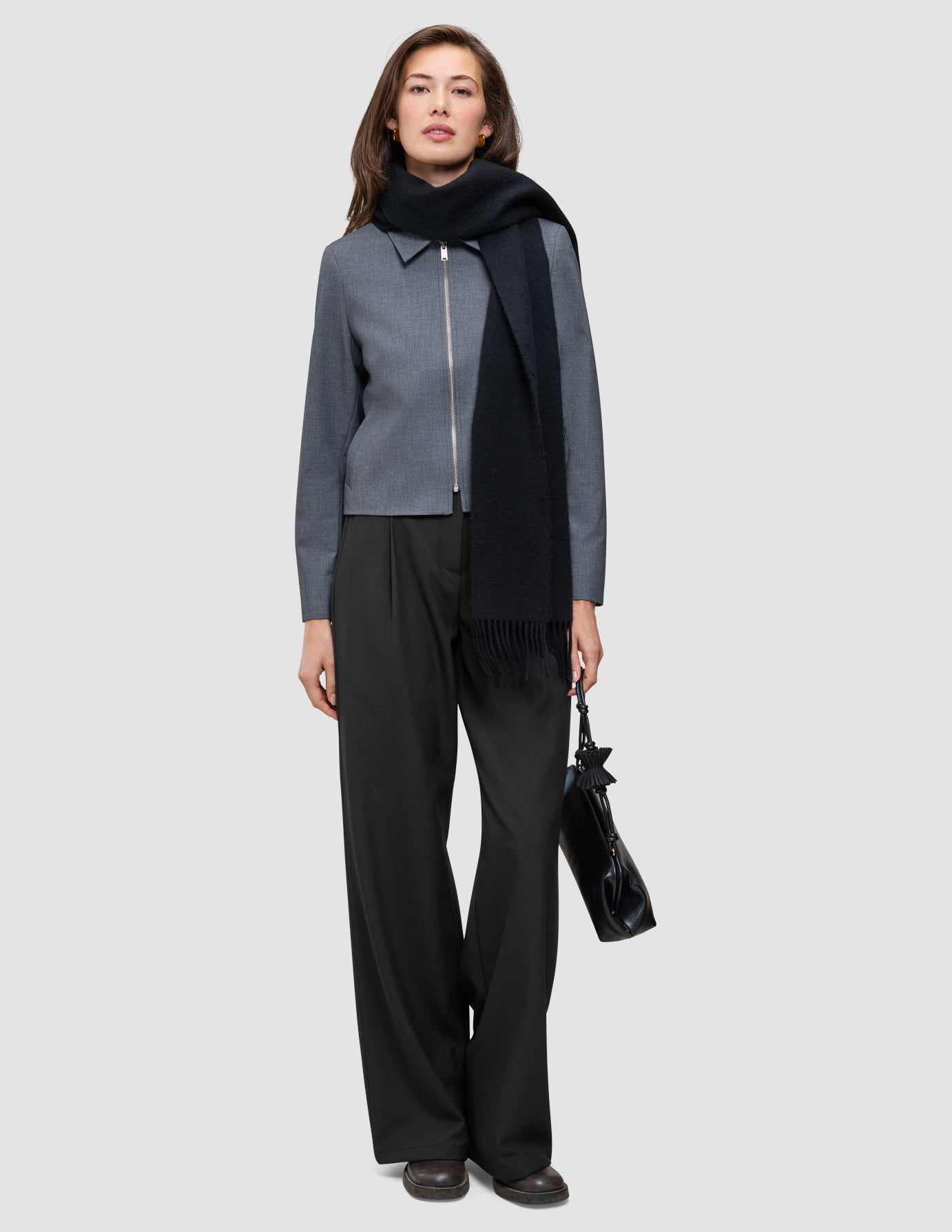 En person klädd i en Essential Short Zip Jacket i mörkgrå melange, en svart halsduk, byxor med vida ben och en svart handväska.