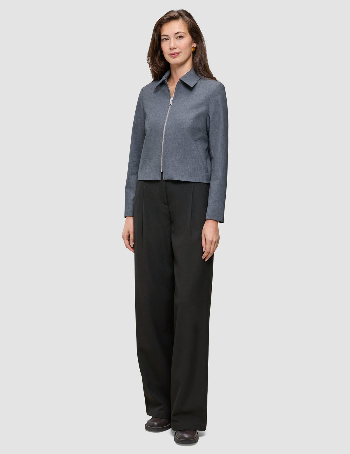 En kvinna klädd i en Essential Short Zip Jacket i mörkgrå melange och svarta byxor i minimalistisk skandinavisk design.