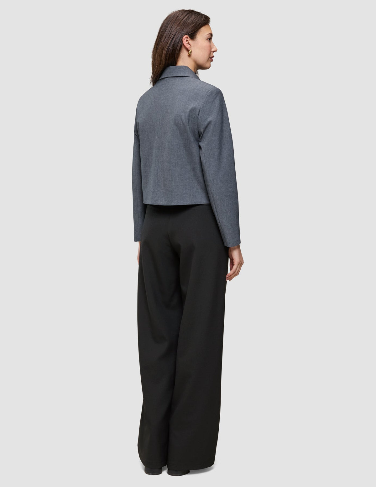 En kvinna sedd bakifrån, klädd i en Essential Short Zip Jacket i mörkgrå melange och svarta byxor.