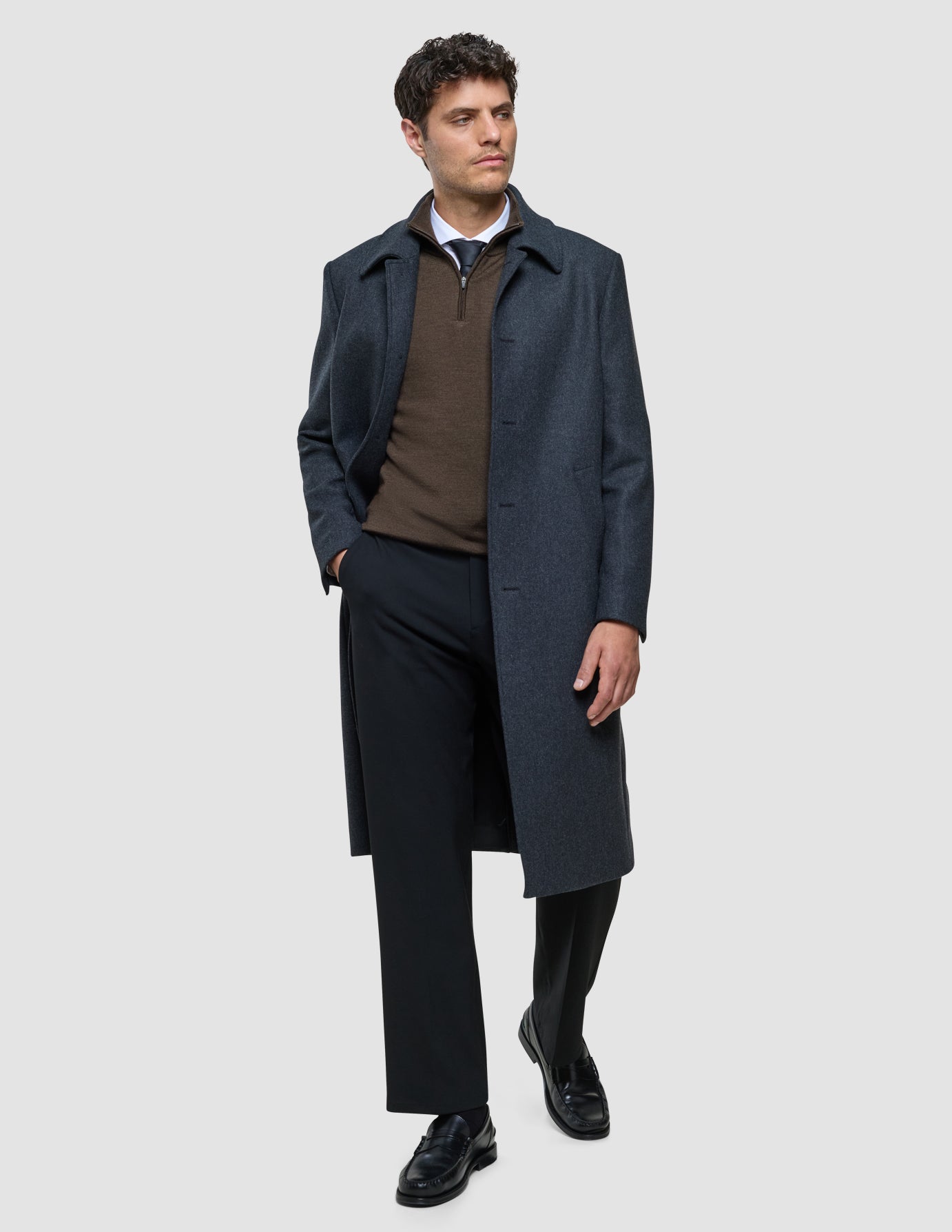 En man bär en mörkgrå Maxi Wool-kappa över en brun tröja, skjorta, slips och svarta byxor.