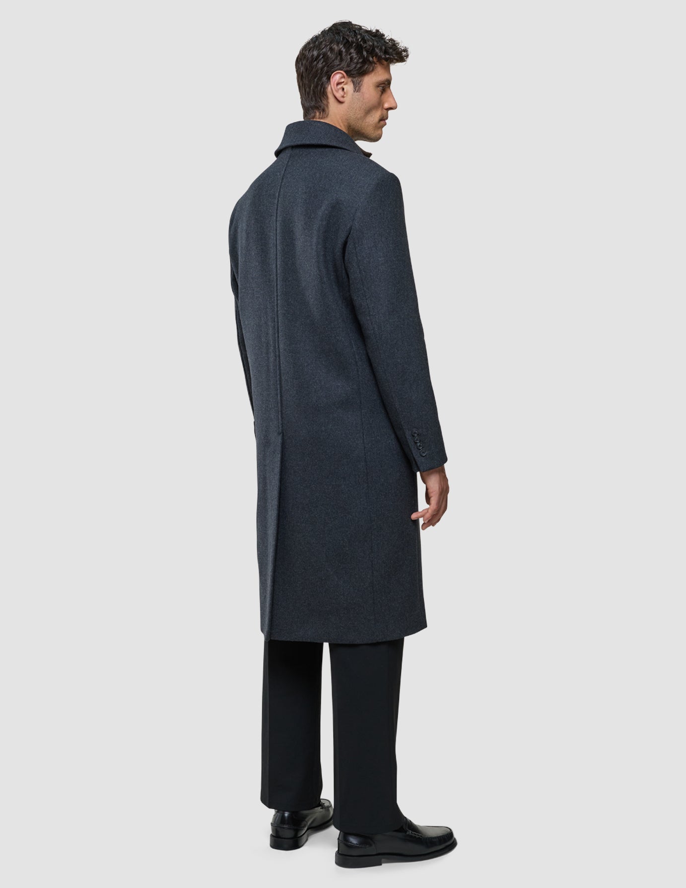 En man står i profil och bär en mörkgrå Maxi Wool Coat Dark Grey i kolgrått som når nedanför knäna.