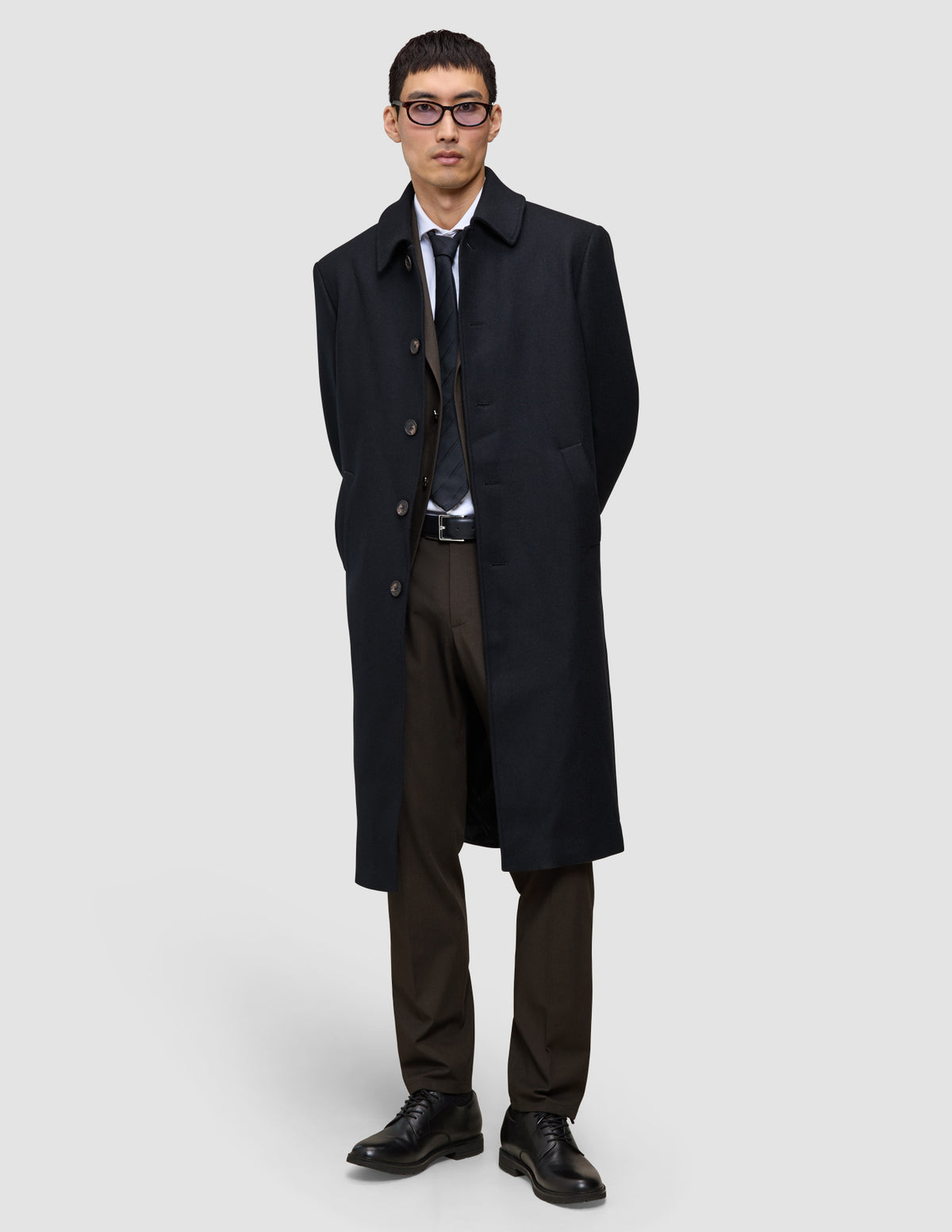En man i svart överrock, kostym, vit skjorta och slips, klädd i byxor av modellen Motion Suit Pants Regular Cognac och svarta kostymskor.