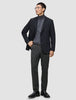 En man klädd i en svart kavaj, grå polotröja, byxor av modellen Motion Suit Pants Regular Pepper Grey, svart bälte och skor.