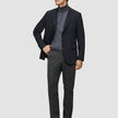 En man klädd i en svart kavaj, grå polotröja, byxor av modellen Motion Suit Pants Regular Pepper Grey, svart bälte och skor.