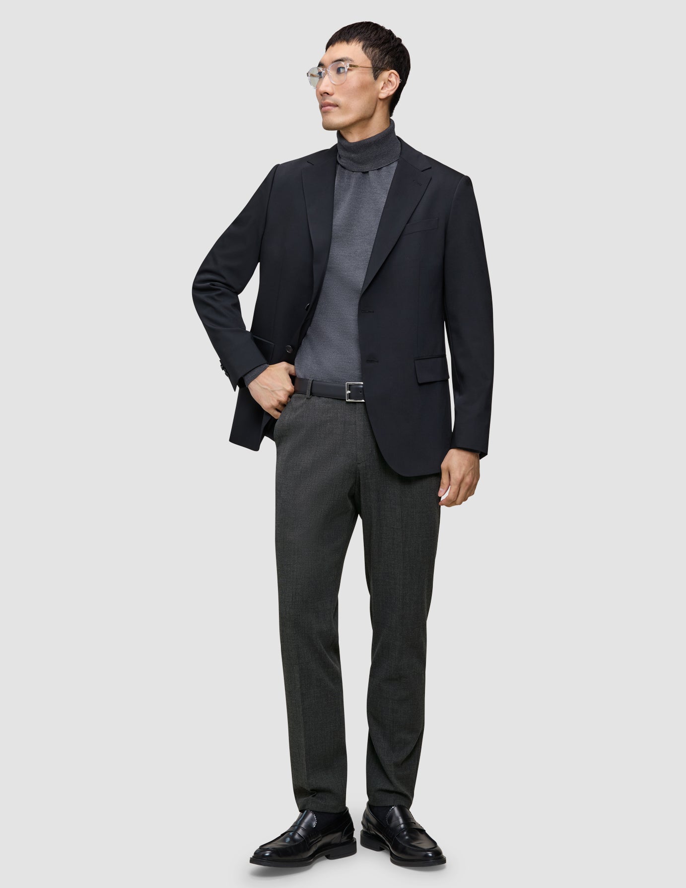En man klädd i en svart kavaj, grå polotröja, byxor av modellen Motion Suit Pants Regular Pepper Grey, svart bälte och skor.