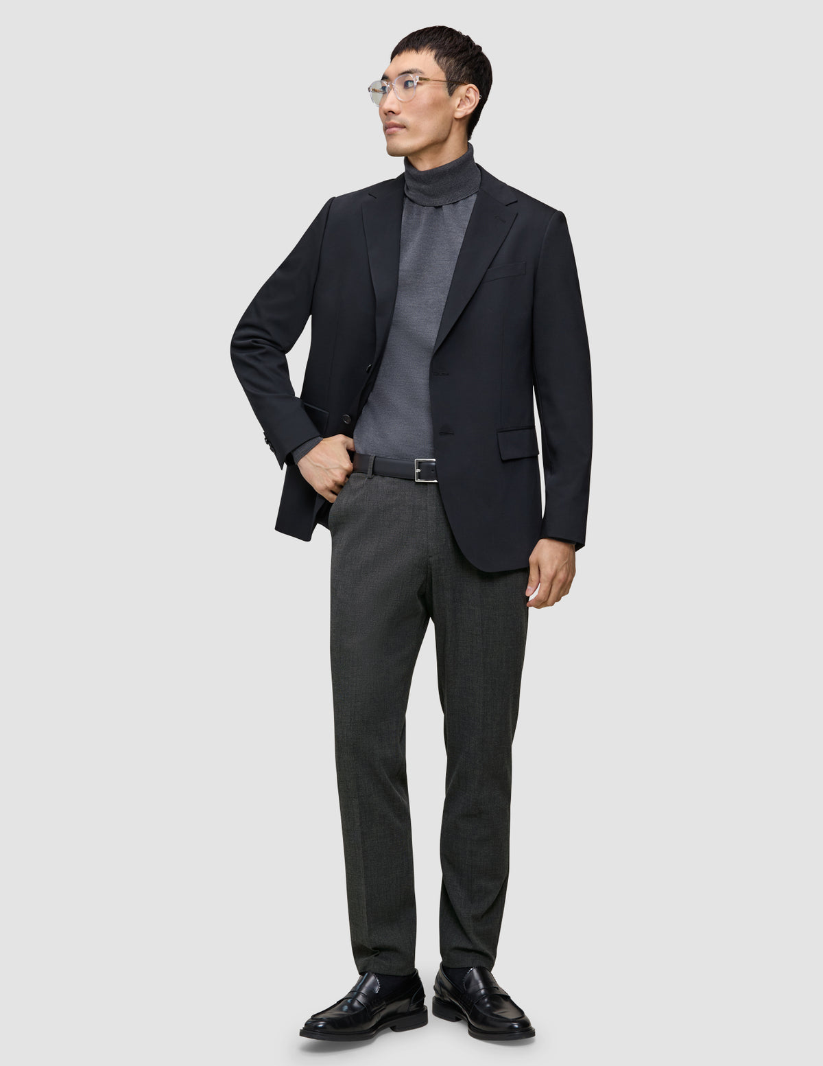 En man klädd i en svart kavaj, grå polotröja, byxor av modellen Motion Suit Pants Regular Pepper Grey, svart bälte och skor.