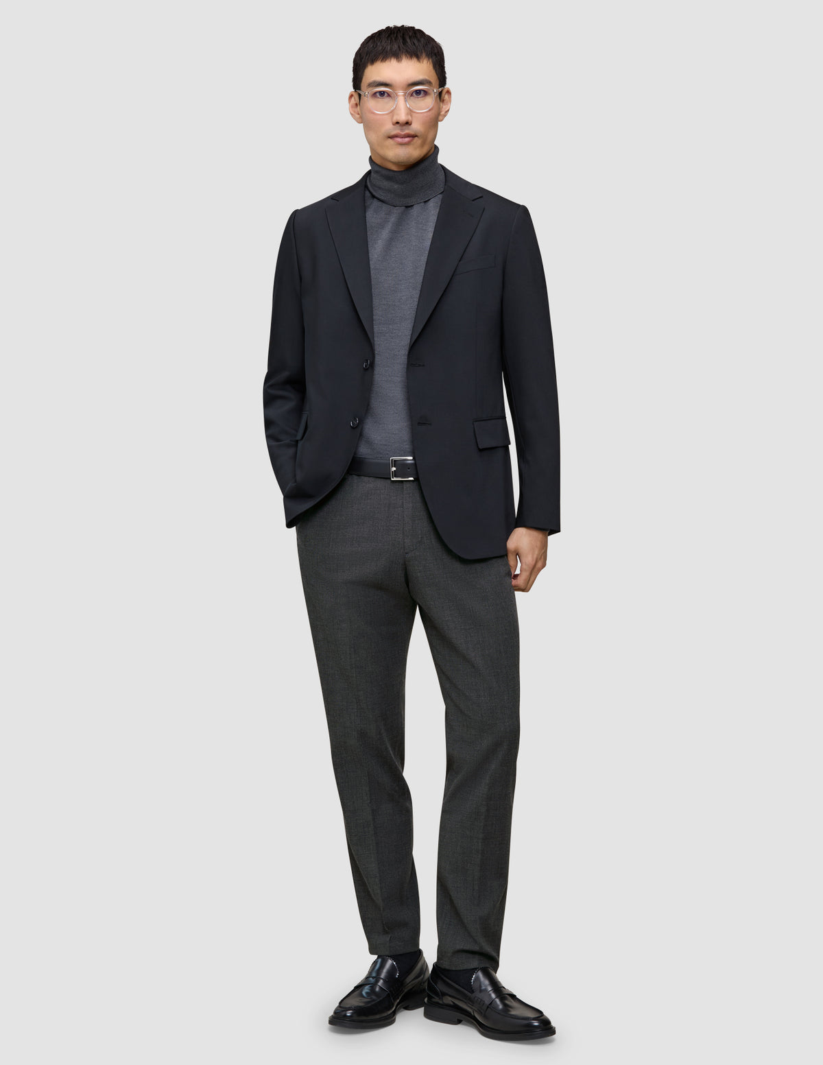 En man i svart kavaj och grå polotröja, klädd i Motion Suit Pants Regular Pepper Grey och svarta loafers.