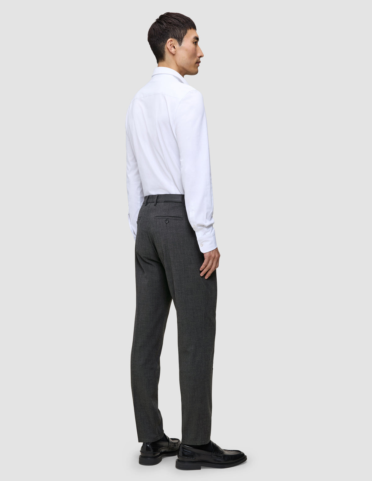 En man sedd bakifrån, klädd i en vit skjorta med knappar, Motion Suit-byxor i storlek Regular i färgen Pepper Grey samt svarta loafers.