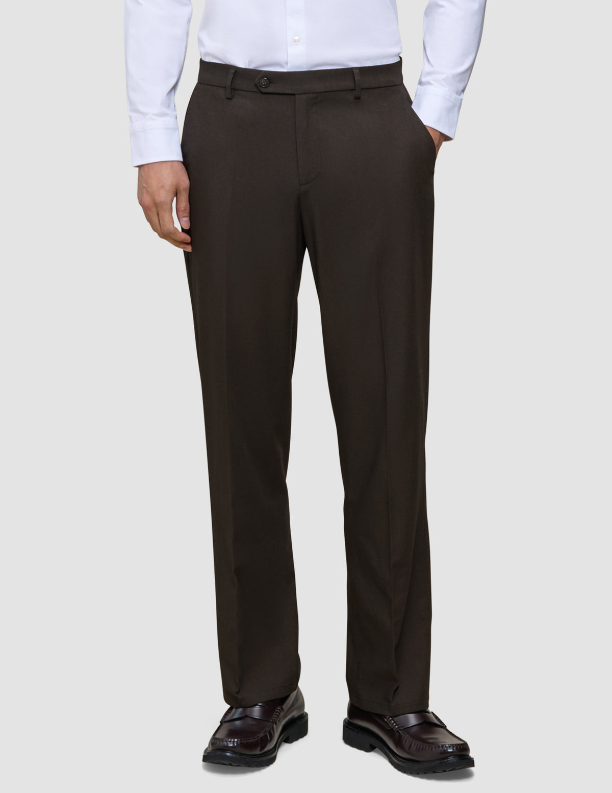 En person bär byxor av modellen Motion Suit Pants Relaxed Cognac, en vit skjorta med knappar och mörka läderskor av typen loafers.