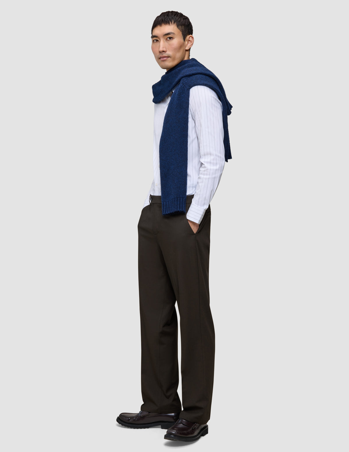 En man bär en vit skjorta med tunna ränder, en blå tröja och byxor av modellen Motion Suit Pants Relaxed Cognac med 360°-stretch.