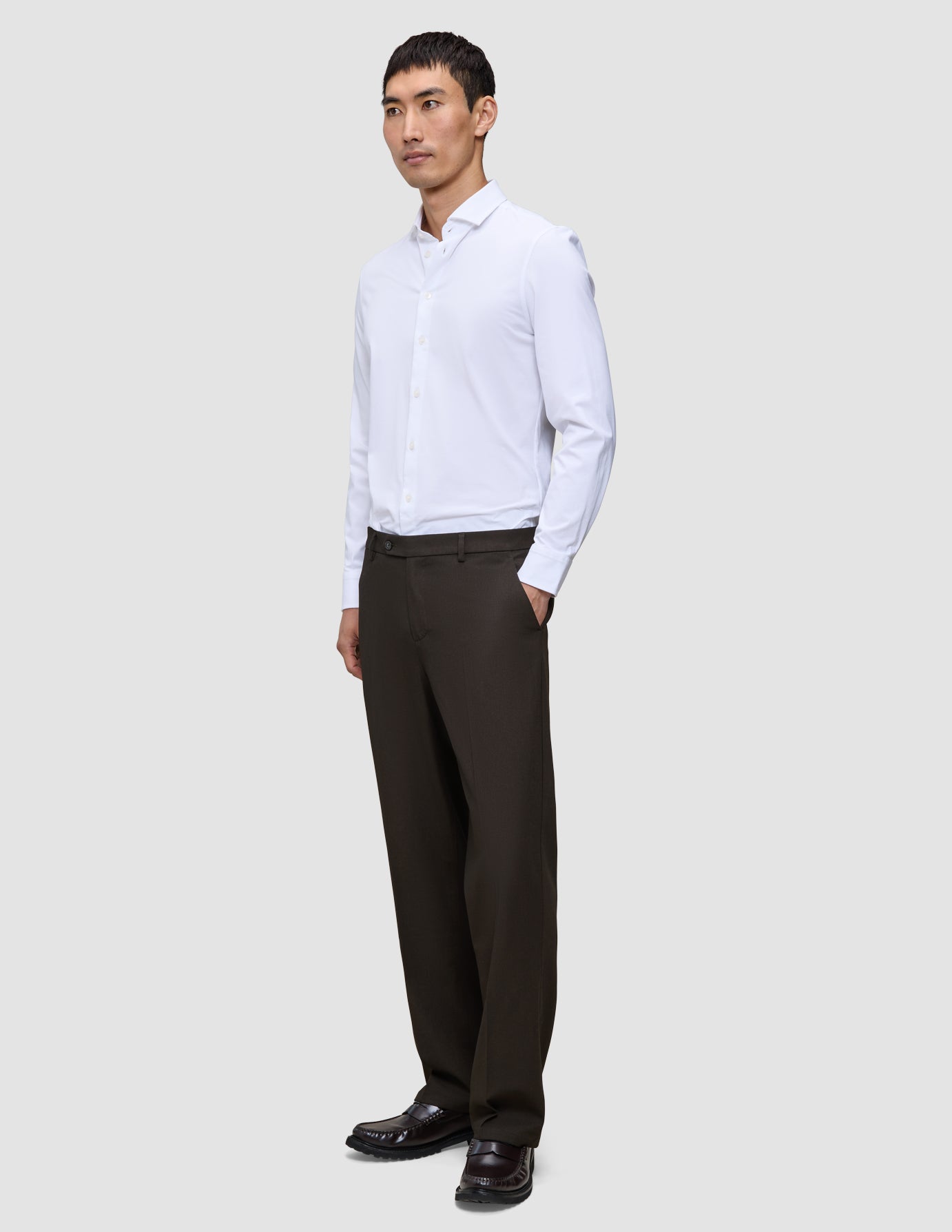 En man i en vit skjorta och ett par Motion Suit-byxor i modell Relaxed i färgen Cognac, med handen i fickan.
